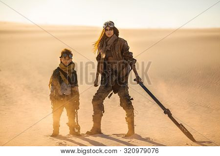 Post Apocalyptic Woman And Boy With Weapons Outdoors. Desert And Dead Wasteland On The Background. Y