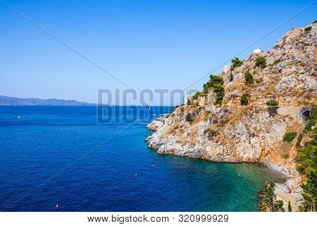 Beautiful Greek Landscape Of Avlaki Beach On Hydra With Tourists Floating In The Sea. Hydra Is One O