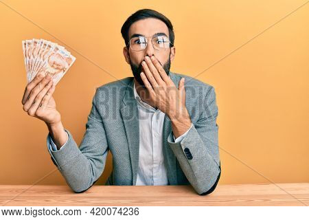 Young hispanic man holding turkish lira banknotes sitting on the table covering mouth with hand, shocked and afraid for mistake. surprised expression 