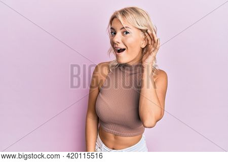 Young blonde girl wearing casual clothes smiling with hand over ear listening and hearing to rumor or gossip. deafness concept. 