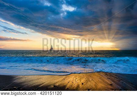 A Sailboat Silhouette Is Sailing Towards The Sun Rays Emanating From The Sunset Sky