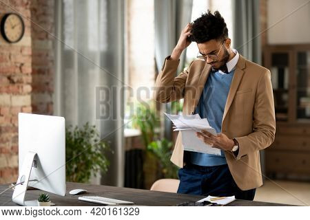 Confused young mixed race man in glasses and jacket standing at desk with computer and scratching head while reading papers