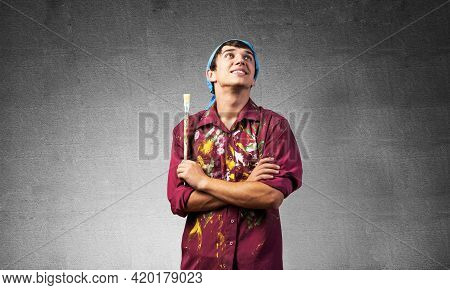 Smiling Young Painter Artist With Folded Arms. Portrait Of Happy Decorator Looking Upwards On Gray W