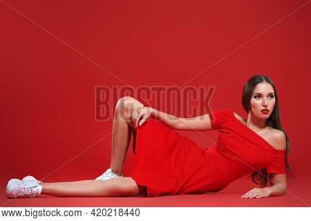 Beautiful brunette girl posing at studio in red dress and white sneakers. Red background. Sport chic style.
