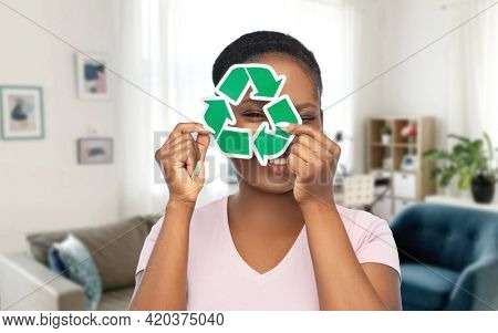 eco living, environment and sustainability concept - portrait of happy smiling young african american woman looking through green recycling sign over home room background