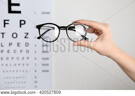 Woman Holding Glasses Against Eye Chart On Light Background, Closeup. Ophthalmologist Prescription