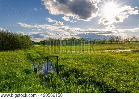 Water Flume With Gate Flowing To Pond On Meadow With Trees And Distant Hill. Czech Spring Landscape