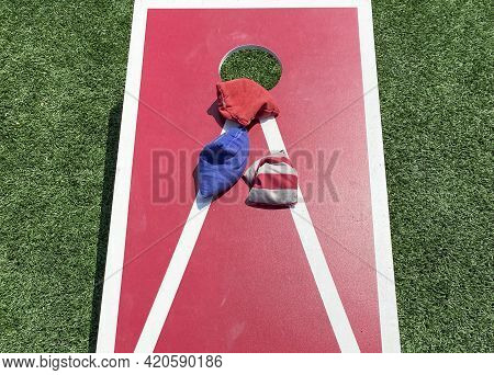A Close Up Of A Red And White Corn Hole Game With Three Bean Bags Close To The Whole.