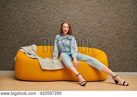 Fashion shot. Beautiful fashion model girl poses in denim jumpsuit on a bright yellow sofa in a modern apartment. Modern interior, furniture.