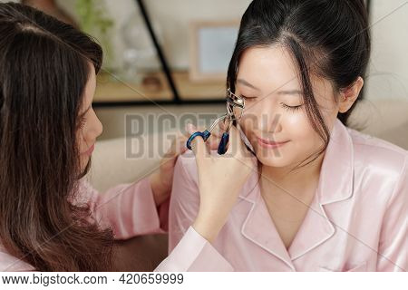 Smiling Young Woman Using Lash Curler When Doing Make-up On Her Best Friend