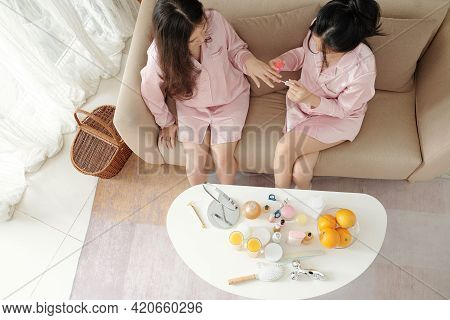 Pretty Friends In Silk Pajamas Sitting On Sofa At Home, Doing Each Other Manicure And Gossiping, Vie