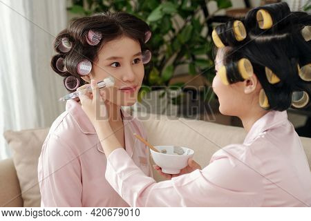 Young Woman Applying Clay Mask On Pretty Face Of Her Best Friend When They Are Enjoying Beauty Party