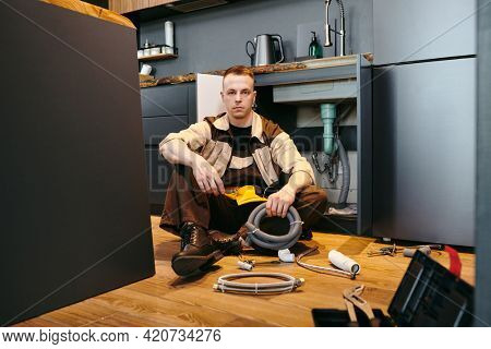 Young plumber in unifrom sitting on the floor by open cabinet while repairing equipmwnt