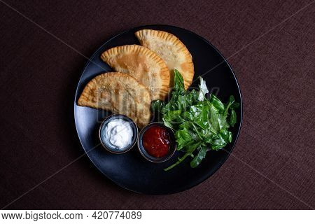 Three Chebureks With Greens On A Black Plate.