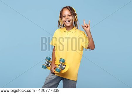 Happy Little Boy With African Dreads With Skateboard On The Blue Background Showing Rock Gesture.