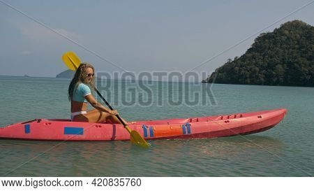 Young Blonde Woman In Blue Swimsuit Rows Pink Plastic Canoe Along Azure Sea Bay Past Island With Pal