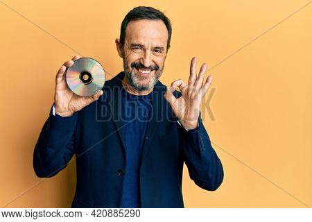 Middle age hispanic man holding compact disc doing ok sign with fingers, smiling friendly gesturing excellent symbol 