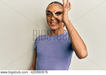 Hispanic man wearing make up and long hair wearing casual t shirt doing ok gesture with hand smiling, eye looking through fingers with happy face. 