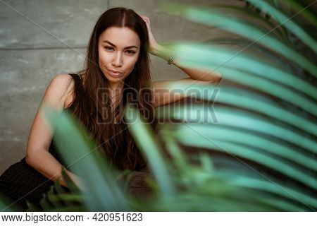 Beautiful Young Woman Closeup Portrait Through Green Leaves Of A Flower On A Gray Wall Background