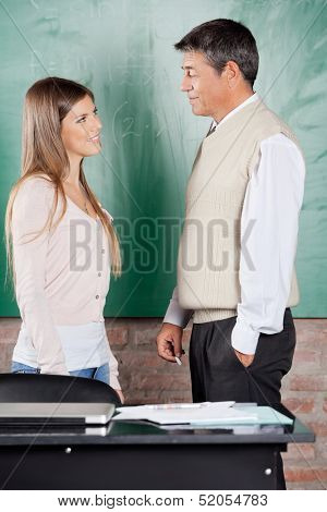 Side view of young college student and teacher looking at each other against greenboard in classroom