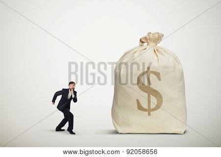 businessman in formal wear showing silent sign and sneaking to the big bag of money over light grey background