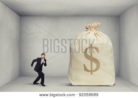 businessman in formal wear showing silent sign and walking on tiptoe to the big bag of money in grey room 