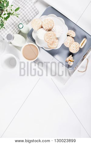 Cookies on white and gray plate with cups of coffee and bottles of milk on white shutters background with green and white flower top view