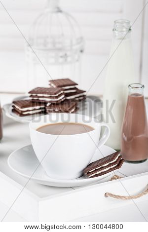 Cup of coffee and few bottles of milk and chocolate milkshakes on a white tray on white shutters background