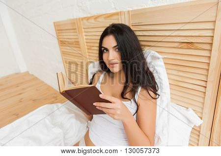 Sexy smart woman  lying on bed and reading book, looking at camera