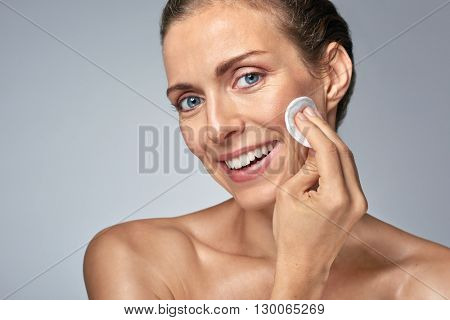 Smiling middle age woman wiping cleaning her face with cotton wool, wellness beauty skin care concept
