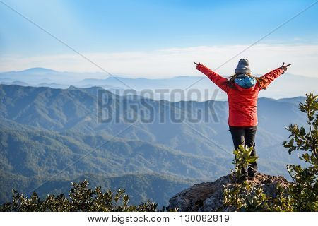 Woman traveler in red coat climb on the rock and upraised arms on top of mountain among beautiful spring landscape - can use for joy or travel concept
