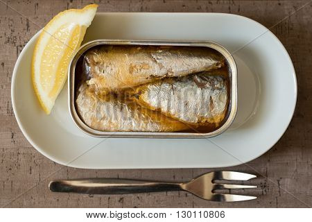 An opened can of sardines or herring and a slice of lemon served on a plate beside a dinner fork