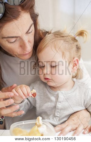 portrait of two years age blonde baby with grey shirt eating from tupperware sitting on legs of mother woman in white table