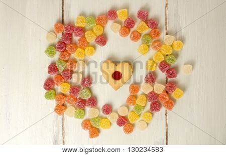 A circle formed by many vibrant gum drops with a heart-shaped tea cookie in the middle shot from above on a light wooden board texture with copyspace