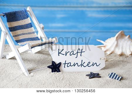 Summer Label With German Text Kraft Tanken Means Relaxation. Blue Wooden Background. Card With Holiday Greetings. Beach Vacation Symbolized By Sand, Deck Chair And Shell.