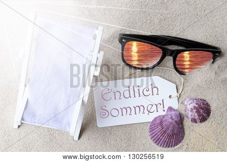 Sunny Summer Label With German Text Endlich Sommer Means Hello Summer. Flat Lay View. Summer Decoration With Deck Chair, Seashells And Sunglasses. Greeting Crad With Sand Background