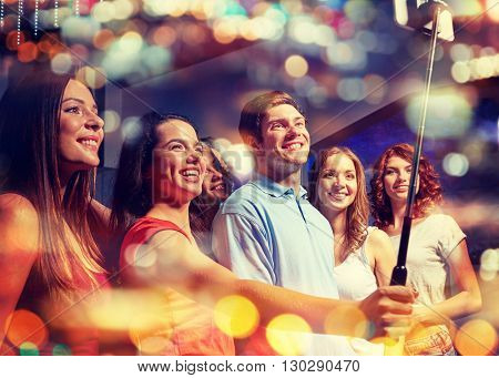 party, technology, nightlife and people concept - smiling friends with smartphone and monopod taking selfie in night club with holidays lights