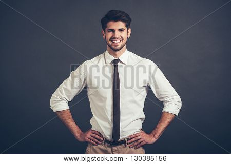 Best smile. Confident young handsome man keeping arms akimbo and looking at camera with smile while standing against grey background