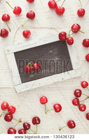 Red cherries scattered on rustic table and heart shaped pair of cherries on empty chalkboard. Overhead view, blank space
