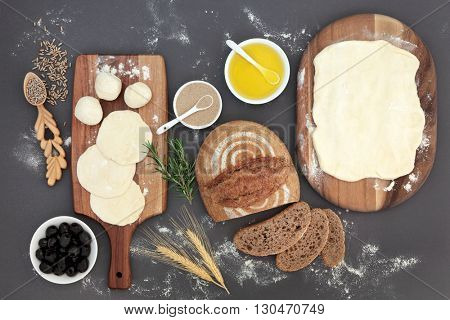 Rustic bread  loaf with baking ingredients of dough on maple boards, olive oil, yeast, herbs, wheat sheaths and rye grain in lovespoon with olives over grey.