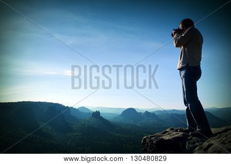 Professional photographer takes photos with big camera on peak of rock. Dreamy misty landscape hot Sun above