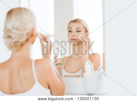beauty, skin care and people concept - smiling young woman applying lotion to cotton disc for washing her face at bathroom