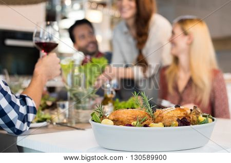 Close up of two roasted chickens with potatoes in a bowl on lunch table. Happy friends having lunch with two grilled chikens with salad. Group of people socializing at home.
