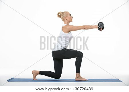 Sportive blonde girl in the sportswear with dumbbells in her hands does split squats on a blue gymnastic mat on the white background in the studio. She wears black pants and white sleeveless t-shirt. She is barefoot. Her hands are raised. She looks forwar