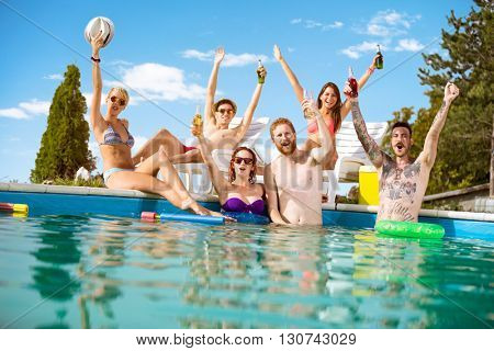Cheerful young people in pool rejoice with beverage in raising hands in pool at summer