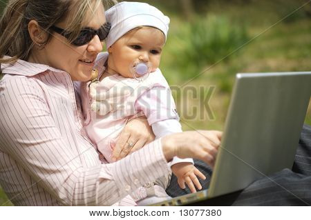 Mutter von einem 9 Monate alten Baby-Mädchen sitzt auf dem Boden im Garten und verwendet einen Laptopcomputer