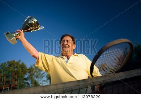 Aktiivinen vanhempi mies hänen 70s poseeraa tenniskentällä kuppia käsissä. Ulkona, auringonvalossa.