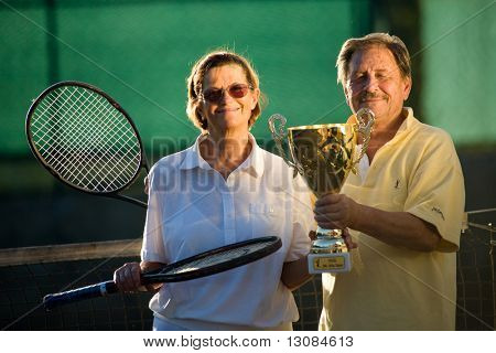 Coppia senior attivo è in posa sul campo da tennis con la racchetta da tennis e la Coppa in mano. All'aperto, sunl