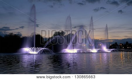 Fountain with bright illumination on the water pond or river with beautiful reflection at the evening or night. Beauty and calm tranquility urban recreational concept.