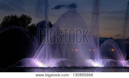 Fountain with bright illumination on the water pond or river with beautiful reflection at the evening or night. Beauty and calm tranquility urban recreational concept.